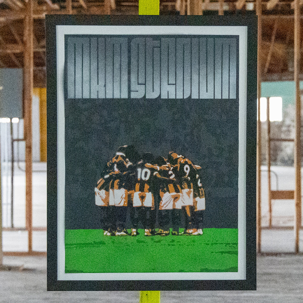 MKM Stadium Hull City hand-sprayed stencil art, amber and black huddle on a green pitch under a charcoal crowd, framed in black, Layered Original edition of 2 by Made By Cito